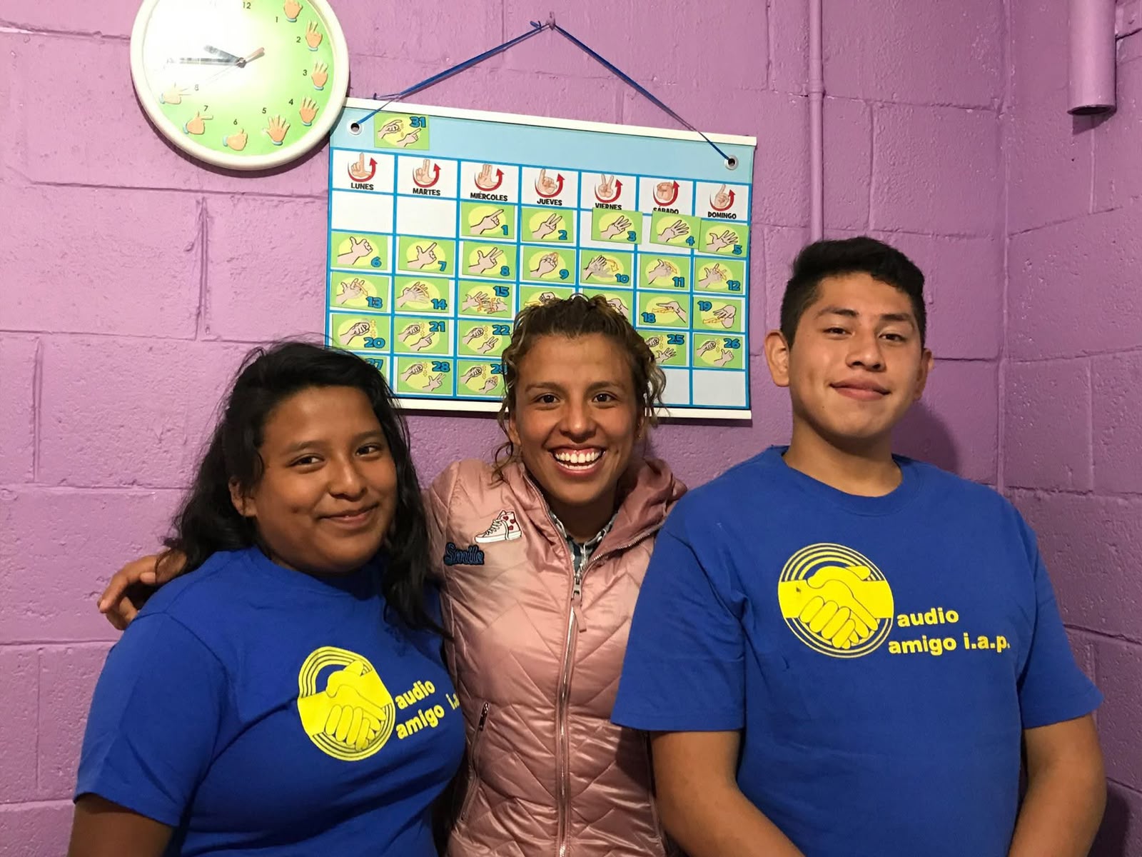 Tres jóvenes de Audio Amigo sonriendo juntos frente a un calendario en lengua de señas